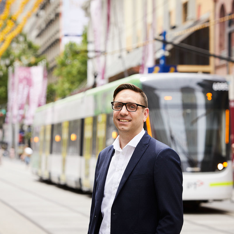 Paul Miziewiczs, RPS Principal Planner stands in busy Bourke Street Mall in Melbourne CBD with a tram behind him.