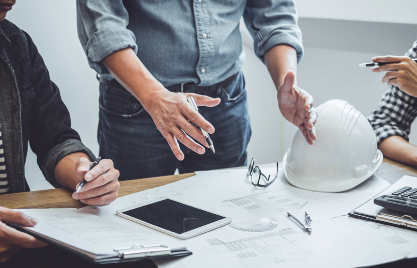 Close up of a development team around a table looking over papers and plans (image shows their mid body and hands). Hard hat, iPad tablet, and calculator also on the table.