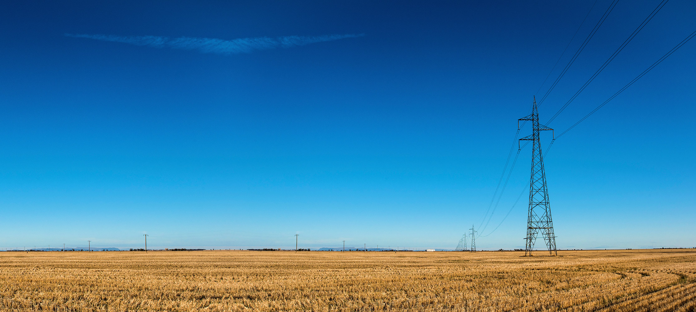Transmission lines extend into the distance across rural farmland in regional Australia. Blue clear skies and recently harvested cropped pasture in the foreground.